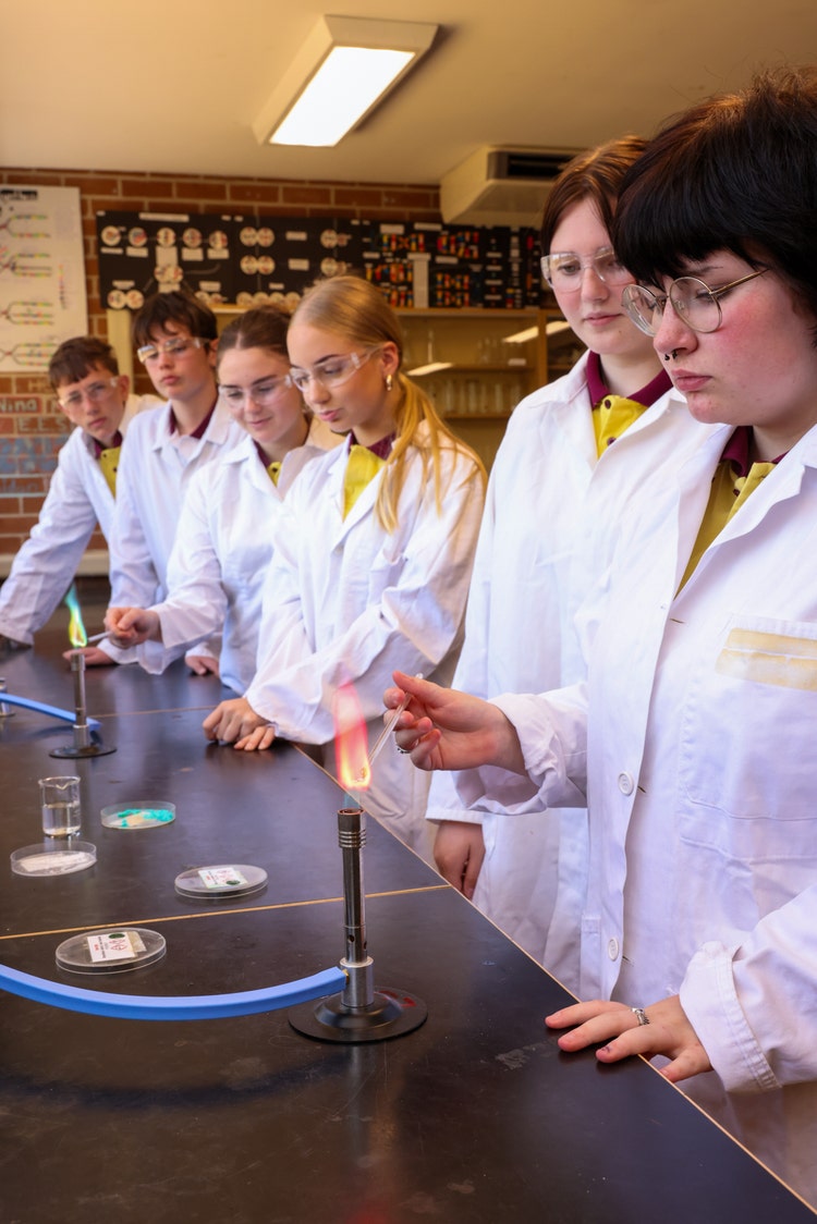 Six students in white lab coats in science lab with two bunsen burners