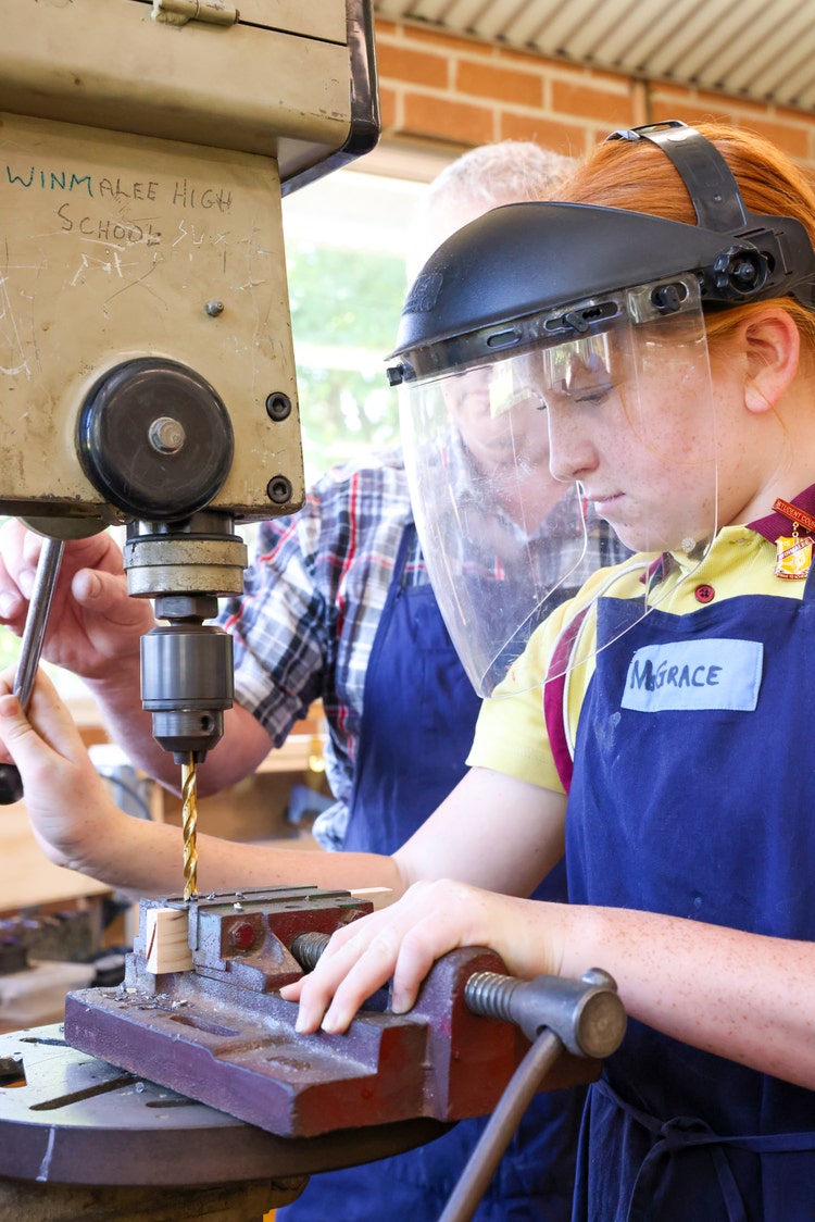 Male teacher and female student wearing safety face shield working on drill press in industrial technology classroom